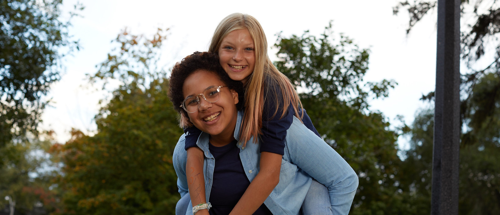 Two Girl Guides ages 12 to 14 outside at a park with one girl giving the other a piggy back ride