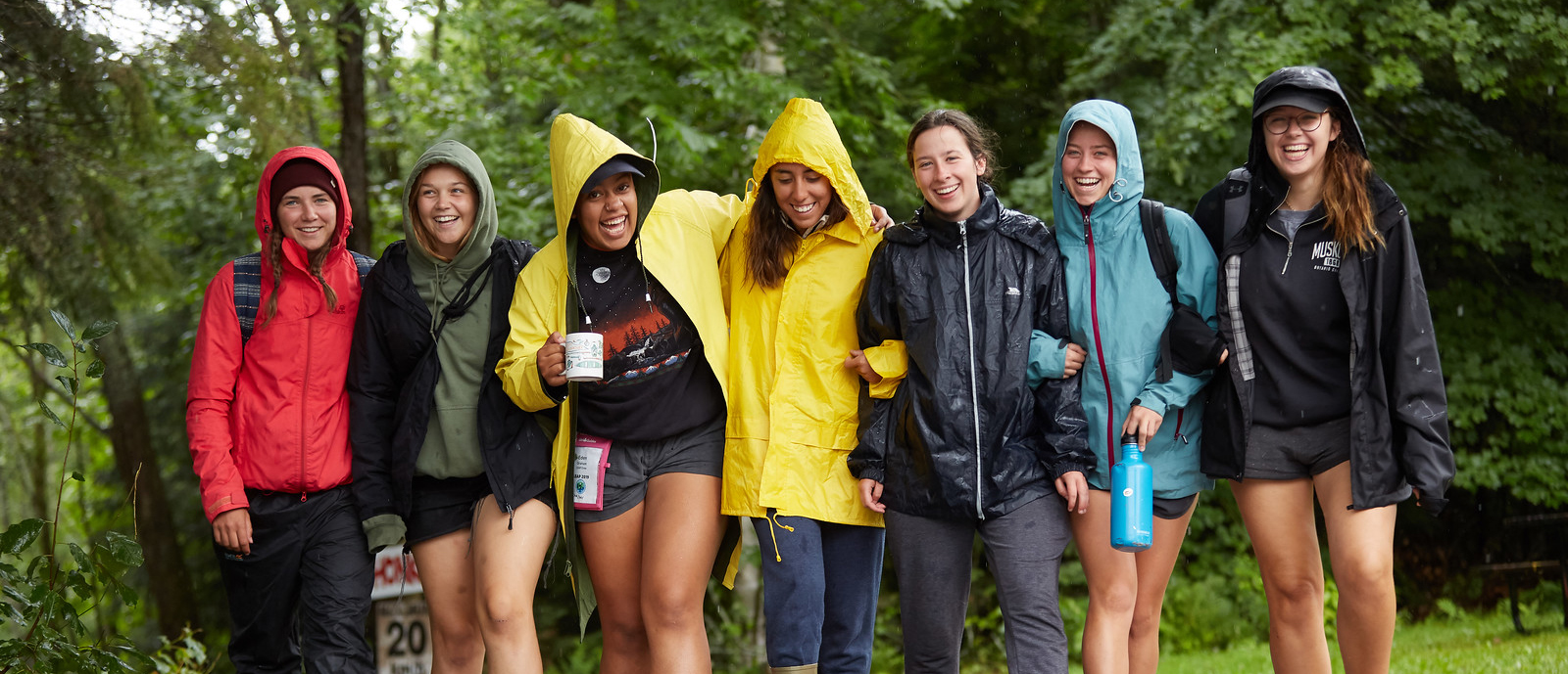 A group of seven Girl Guides ages 15 to 17 smiling and walking together in the woods while it’s raining. They are wearing rain jackets, some with hoods up.